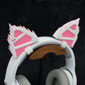 May include: White headphones with pink cat ears attached to the headband. The headphones are on a wooden stand.