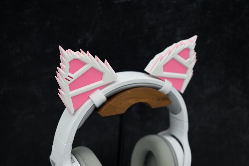 May include: White headphones with pink cat ears attached to the headband. The headphones are on a wooden stand.