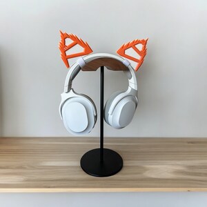 May include: White wireless headphones with orange fox ear covers on a black stand. The headphones are on a wooden surface.