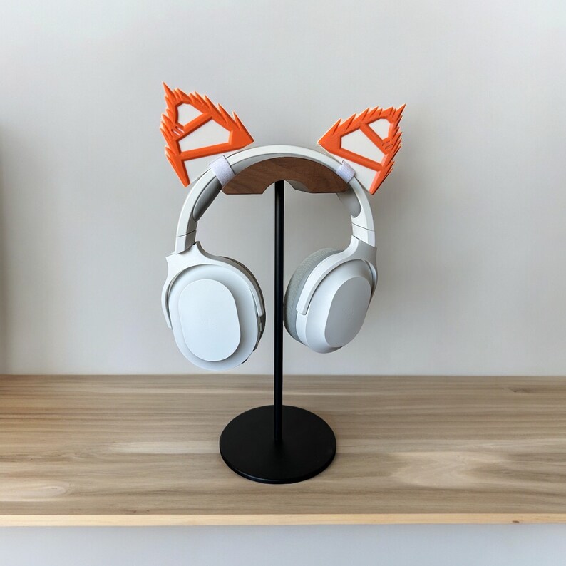May include: White wireless headphones with orange fox ear covers on a black stand. The headphones are on a wooden surface.