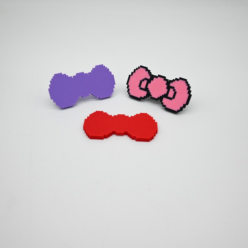 May include: Three pixelated bow shapes made of plastic. One is purple, one is red, and one is pink with a black outline.