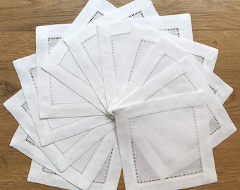 Op de afbeelding: Een set van 12 witte linnen servetten met een delicate geborduurde rand. De servetten zijn in een waaier gelegd, waardoor de ingewikkelde details zichtbaar worden.