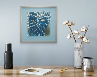 May include: A framed abstract art piece with a blue and gray radial design. The artwork is displayed on a light blue wall, alongside a vase of cotton stems, a dark gray bottle, and a magazine. The overall aesthetic is modern and minimalist.