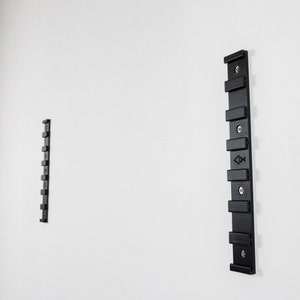 Puede incluir: Un soporte de pared negro con una serie de ranuras para colgar objetos. El soporte está fijado a una pared blanca.