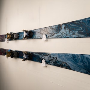 Puede incluir: Dos pares de esquís montados en una pared blanca. Los esquís tienen un diseño veteado azul y negro con arte lineal de montañas. Las fijaciones son negras y doradas. Soportes blancos para esquís montados en la pared.