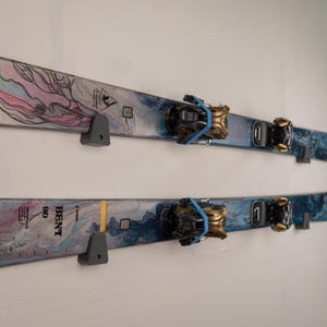 May include: Two pairs of skis mounted on a white wall. The skis have a blue and white marbled design with pink and purple accents. The bindings are black and gold. The word "BENT" is visible on one ski.