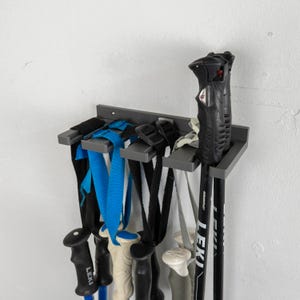 May include: A grey metal ski pole rack fixed to a white wall. The rack displays several pairs of ski poles with black, blue, and white straps. The ski poles are black with the brand name "LEKI" visible.