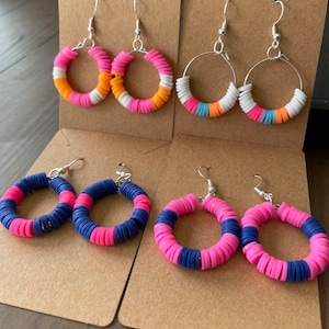May include: Four pairs of hoop earrings with silver hooks. The earrings feature colorful, stacked polymer clay beads in various color combinations: pink, orange, white, and blue. The earrings are displayed on brown cardstock.