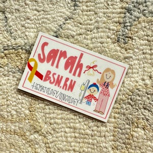 May include: A pink and white card with the name "Sarah BSN, RN" and the words "Hematology/Oncology". The card features a cartoon illustration of a woman with a stethoscope and a child with an IV drip.