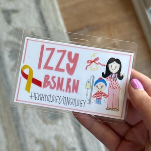 May include: A pink and white ID badge with the name "Izzy BSN, RN" and the text "Hematology/Oncology". The badge features a cartoon illustration of a woman in a pink dress and a child in a red and white striped shirt.