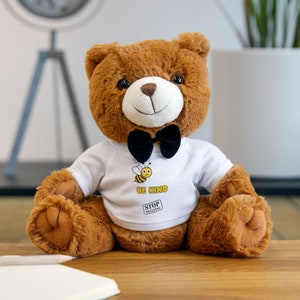 May include: A brown teddy bear wearing a white shirt with a bee and the text "Be Kind" and a black bow tie. The shirt also has a red square with the text "Stop Bullying".
