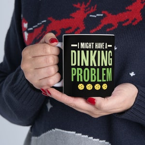 May include: Black coffee mug with white text and a green background. The text reads "I might have a dinking problem" with four yellow pickleball icons at the bottom.