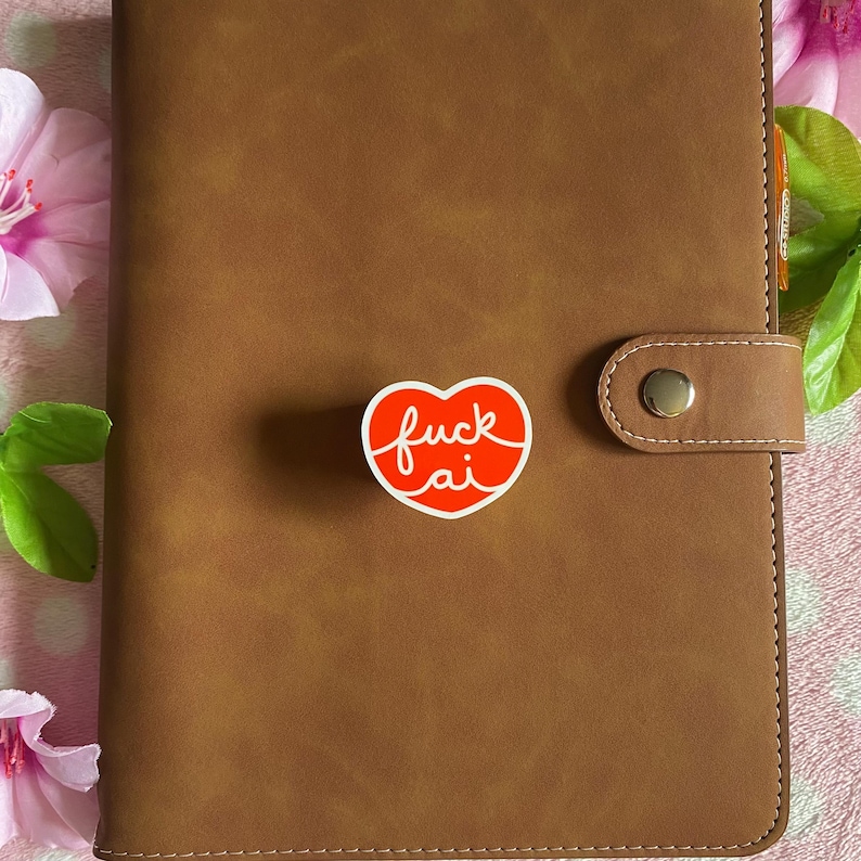 May include: A brown leather-bound planner with a snap closure. A red heart-shaped sticker with the words "fuck ai" in white script is affixed to the cover. The planner is surrounded by pink flowers.