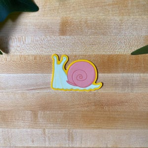 May include: A die-cut sticker featuring a cartoon snail with a pale blue body and a rounded pink shell. The snail has two eye stalks and a distinct yellow outline surrounding the entire shape, creating a bold and clear graphic design.