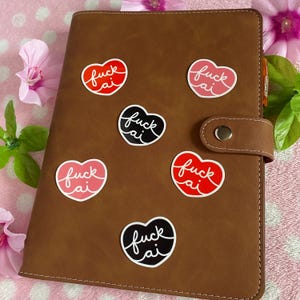 May include: A brown leather planner with several heart-shaped stickers. Each sticker has the words "fuck ai" written in a cursive font. The stickers are in various colors, including red, pink, and black. The planner is closed and has a button closure.