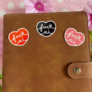May include: Three heart-shaped stickers with the text "fuck ai". One sticker is red with an orange outline, one is black with a white outline, and the last is pink with a white outline. The stickers are on a brown leather-like planner.