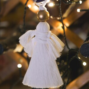 Op de afbeelding: Witte kwastversiering met een gouden kraal aan de bovenkant. De versiering hangt aan een lichtsnoer.