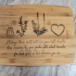 Op de afbeelding: Een rechthoekige bamboe snijplank met lasergegraveerde illustraties van een zoutvaatje, een takje rozemarijn, een lavendelplant en een hart. De tekst luidt: Always throw spilt salt over your left shoulder keep rosemary by your garder gate plant lavender for luck fall in love wherever you can.