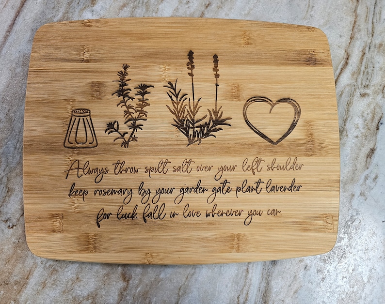 Op de afbeelding: Een rechthoekige bamboe snijplank met lasergegraveerde illustraties van een zoutvaatje, een takje rozemarijn, een lavendelplant en een hart. De tekst luidt: Always throw spilt salt over your left shoulder keep rosemary by your garder gate plant lavender for luck fall in love wherever you can.