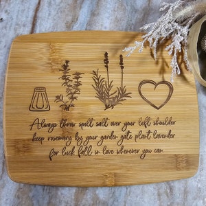Op de afbeelding: Een rechthoekige bamboe snijplank met in laser gegraveerde illustraties van een zoutvaatje, een takje rozemarijn, een lavendelplant en een hart. De tekst luidt: Always throw spill salt over your left shoulder keep rosemary by your garden gate plant lavender for luck fall in love wherever you can.