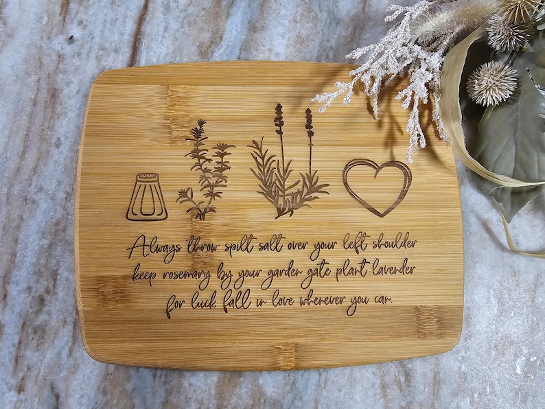 Op de afbeelding: Een rechthoekige bamboe snijplank met in laser gegraveerde illustraties van een zoutvaatje, een takje rozemarijn, een lavendelplant en een hart. De tekst luidt: Always throw spill salt over your left shoulder keep rosemary by your garden gate plant lavender for luck fall in love wherever you can.
