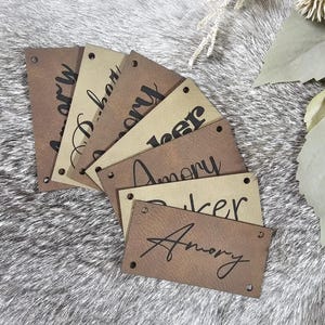 May include: A set of six brown leather tags with the name "Amory" written in black lettering. The tags have holes punched in the corners and are arranged in a fan shape.