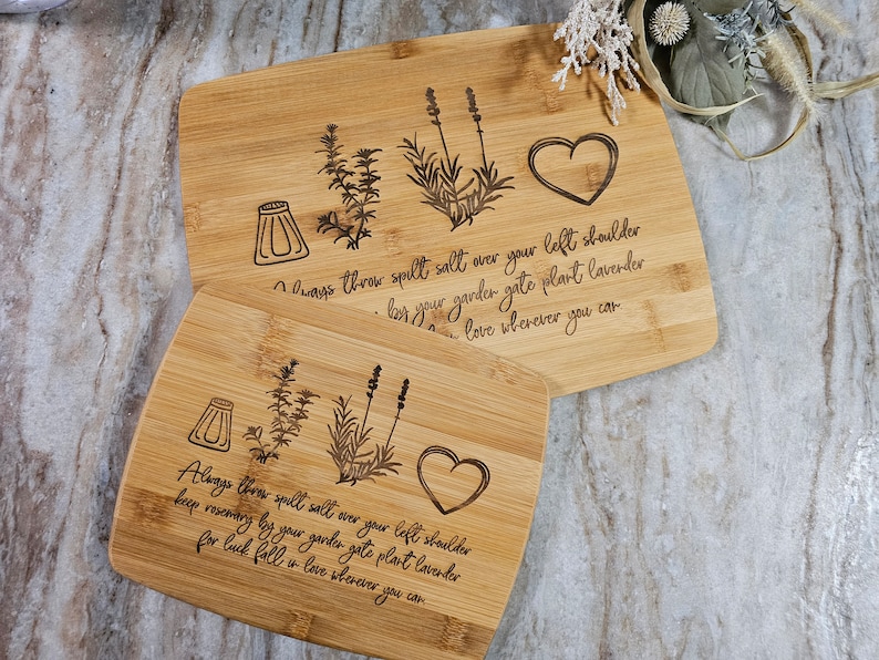 Op de afbeelding: Twee rechthoekige bamboe snijplanken met lasergegraveerde illustraties van een zoutvaatje, een takje rozemarijn, een lavendelplant en een hart. De tekst luidt: Always throw spelt salt over your left shoulder, keep rosemary by your garden gate, plant lavender for luck, fall in love wherever you can.
