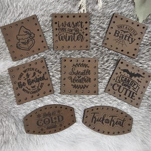 May include: Eight brown leather patches with various Halloween and winter-themed sayings. The patches have a distressed look and are decorated with small silver studs. The sayings include "I wasn't made for winter", "Just a little batty", "This witch can be bribed with chocolate", "Sweater weather", "Candy corn cutie", "Baby it's cold outside", and "Trick or treat".