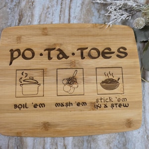 May include: A rectangular bamboo cutting board with the word "po-ta-toes" engraved at the top. Below are three illustrated steps: boil 'em, mash 'em, and stick 'em in a stew. The board has a natural wood grain.