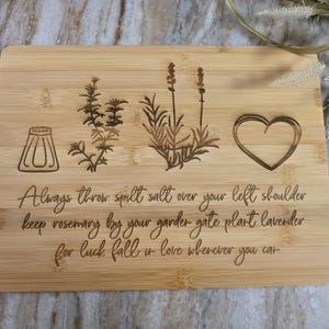 Op de afbeelding: Een rechthoekige bamboe snijplank met lasergegraveerde illustraties van een zoutvaatje, een takje rozemarijn, een lavendelplant en een hart. De tekst luidt: Always throw spill salt over your left shoulder keep rosemary by your garden gate plant lavender for luck fall in love wherever you can.