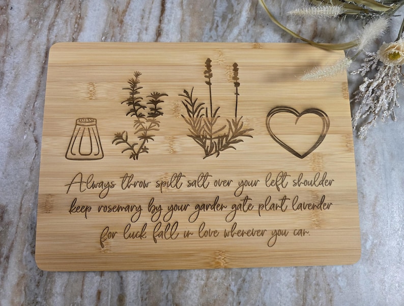 Op de afbeelding: Een rechthoekige bamboe snijplank met lasergegraveerde illustraties van een zoutvaatje, een takje rozemarijn, een lavendelplant en een hart. De tekst luidt: Always throw spill salt over your left shoulder keep rosemary by your garden gate plant lavender for luck fall in love wherever you can.