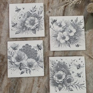 May include: Four square ceramic coasters with a floral and bee design. Each coaster features a different arrangement of flowers, bees, and honeycomb patterns in shades of gray on a white background. The coasters are arranged on a marble surface.