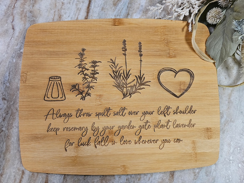 Op de afbeelding: Een rechthoekige bamboe snijplank met lasergegraveerde illustraties van een zoutvaatje, een takje rozemarijn, een lavendelstengel en een hart. De tekst luidt: Always throw spilt salt over your left shoulder keep rosemary by your garden gate plant lavender for luck fall in love wherever you can.