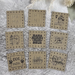 May include: Nine square, tan-colored coasters with various black text designs. The coasters include phrases like "Mama," "Mini," "It's all a bunch of hocus pocus," "Dog mom," and "Drink Happy thoughts." Each coaster has a dotted border.