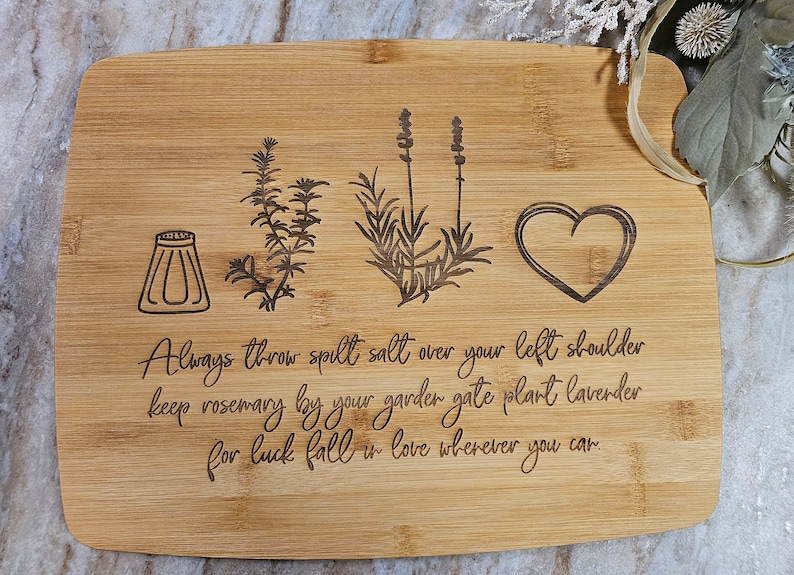 Op de afbeelding: Een rechthoekige bamboe snijplank met lasergegraveerde illustraties van een zoutvaatje, een takje rozemarijn, een lavendelstengel en een hart. De tekst luidt: Always throw spilt salt over your left shoulder keep rosemary by your garden gate plant lavender for luck fall in love wherever you can.