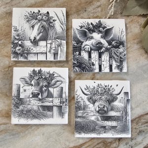 May include: Set of four black and white ceramic coasters featuring farm animals with floral wreaths. Each coaster depicts a different animal: a horse, a pig, a cow, and a Highland cow. The coasters are square and have a rustic, farmhouse style.
