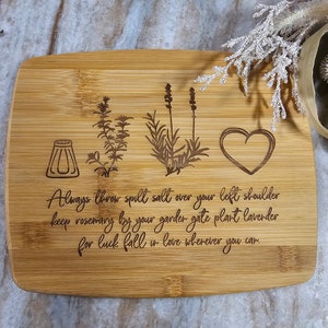 Op de afbeelding: Een rechthoekige bamboe snijplank met lasergegraveerde illustraties van een zoutvaatje, een takje rozemarijn, een lavendelplant en een hart. De tekst luidt: Always throw spill salt over your left shoulder keep rosemary by your garder gate plant lavender for luck fall in love wherever you car.