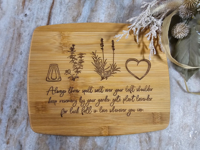 Op de afbeelding: Een rechthoekige bamboe snijplank met lasergegraveerde illustraties van een zoutvaatje, een takje rozemarijn, een lavendelplant en een hart. De tekst luidt: Always throw spill salt over your left shoulder keep rosemary by your garder gate plant lavender for luck fall in love wherever you car.