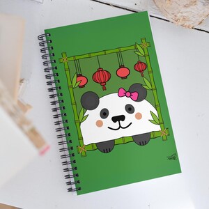 Pode incluir: Um caderno espiral verde com uma ilustração de um panda. O panda está usando um laço rosa e está espiando por trás de uma moldura de bambu. Lanternas vermelhas penduradas no bambu.