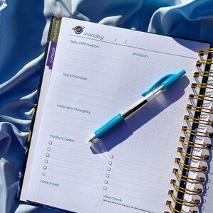 Pode incluir: Um planejador aberto em uma p&aacute;gina de segunda-feira com se&ccedil;&otilde;es para afirma&ccedil;&otilde;es di&aacute;rias e tarefas. Uma caneta azul est&aacute; sobre a p&aacute;gina. O caderno espiralado tem uma capa azul e dourada.
