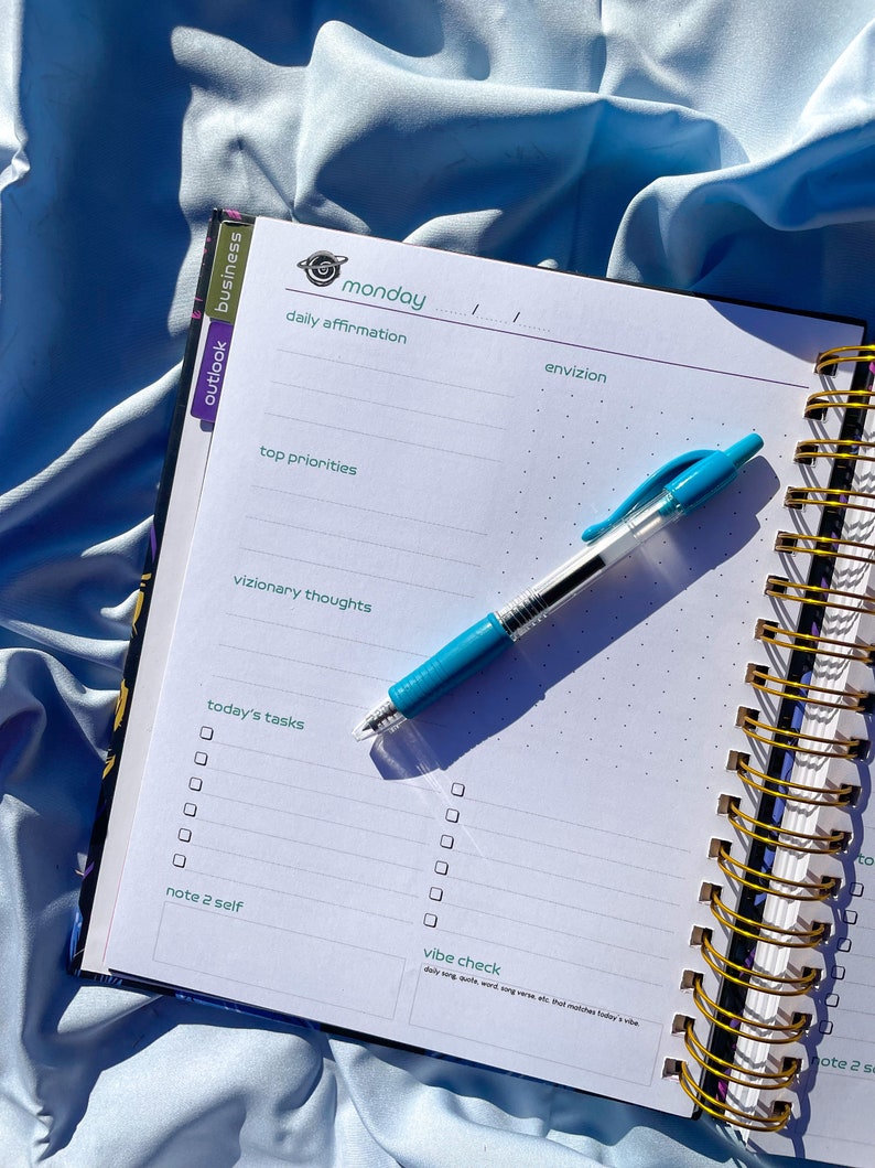 Pode incluir: Um planejador aberto em uma p&aacute;gina de segunda-feira com se&ccedil;&otilde;es para afirma&ccedil;&otilde;es di&aacute;rias e tarefas. Uma caneta azul est&aacute; sobre a p&aacute;gina. O caderno espiralado tem uma capa azul e dourada.