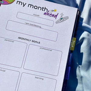 Pode incluir: Agenda aberta com o texto "my month ahead" e se&ccedil;&otilde;es para metas mensais. O planejador tem um esquema de cores roxo e branco com uma ilustra&ccedil;&atilde;o com tema espacial. As abas laterais s&atilde;o rotuladas como "focus", "direction" e "structure".