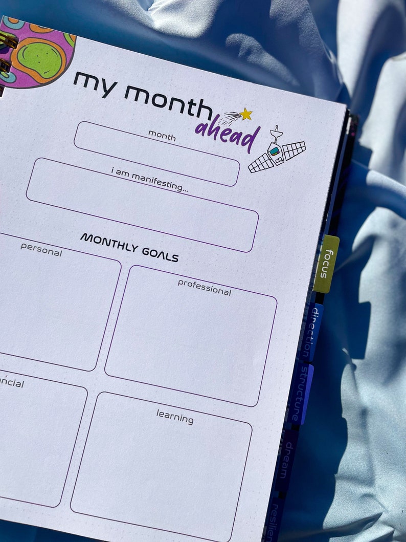 Pode incluir: Agenda aberta com o texto "my month ahead" e se&ccedil;&otilde;es para metas mensais. O planejador tem um esquema de cores roxo e branco com uma ilustra&ccedil;&atilde;o com tema espacial. As abas laterais s&atilde;o rotuladas como "focus", "direction" e "structure".