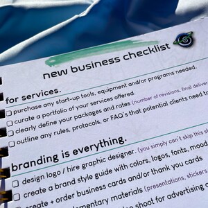 Pode incluir: Um caderno espiralado com uma lista de verifica&ccedil;&atilde;o intitulada "New Business Checklist". A lista de verifica&ccedil;&atilde;o inclui se&ccedil;&otilde;es para servi&ccedil;os e branding, com caixas de sele&ccedil;&atilde;o ao lado de cada item. O caderno est&aacute; em uma superf&iacute;cie azul.