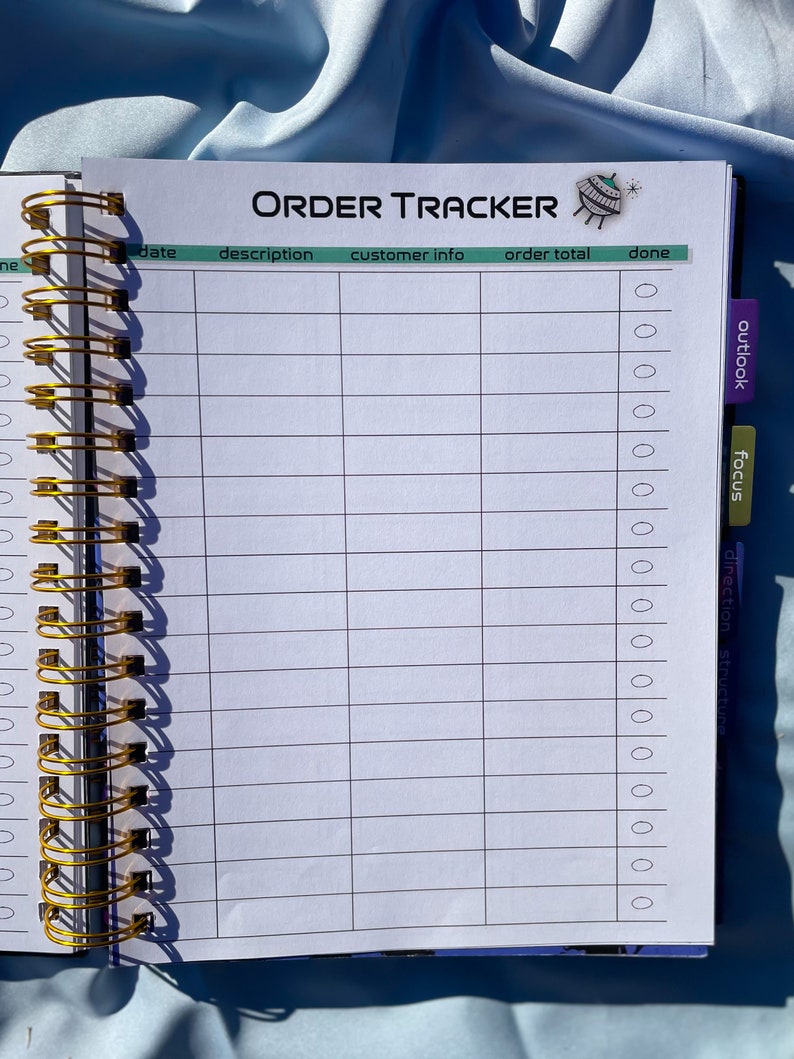 Pode incluir: Um caderno aberto com espiral com as palavras "ORDER TRACKER" impressas no topo. O caderno tem uma encaderna&ccedil;&atilde;o em espiral dourada e cont&eacute;m p&aacute;ginas pautadas com colunas para detalhes do pedido. Abas rotuladas como "outlook" e "focus" s&atilde;o vis&iacute;veis.