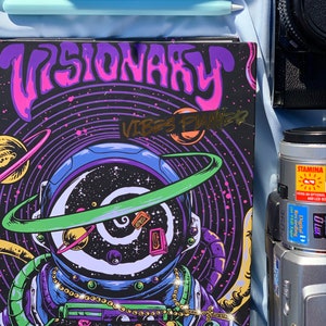 Pode incluir: Um planejador com o texto "VISIONARY VIBES PLANNER" em um design psicod&eacute;lico com um astronauta, planetas e estrelas. O planejador &eacute; preto com detalhes em roxo, rosa, verde e dourado. Um telefone azul, uma caneta e uma c&acirc;mera tamb&eacute;m s&atilde;o vis&iacute;veis.