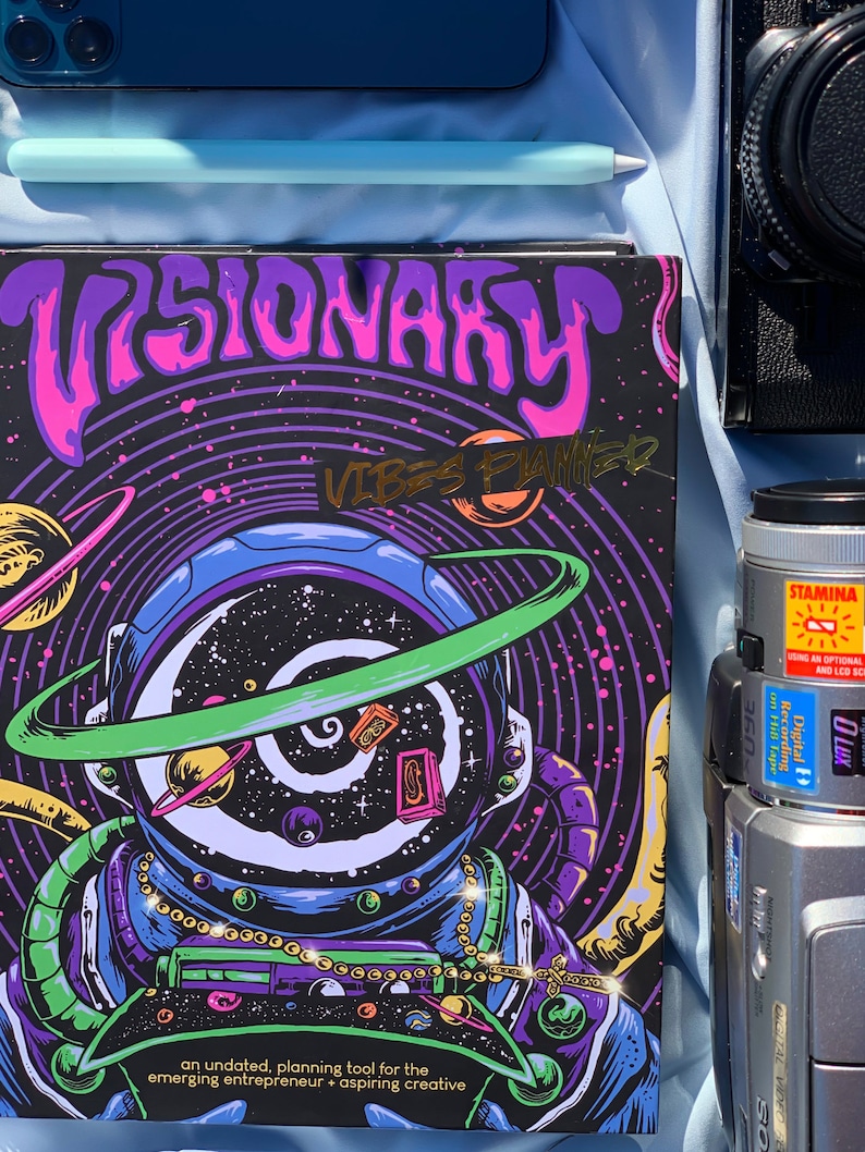 Pode incluir: Um planejador com o texto "VISIONARY VIBES PLANNER" em um design psicod&eacute;lico com um astronauta, planetas e estrelas. O planejador &eacute; preto com detalhes em roxo, rosa, verde e dourado. Um telefone azul, uma caneta e uma c&acirc;mera tamb&eacute;m s&atilde;o vis&iacute;veis.