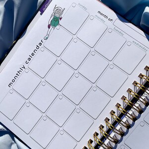 Pode incluir: Um calend&aacute;rio mensal com encaderna&ccedil;&atilde;o em espiral com interior branco e encaderna&ccedil;&atilde;o em metal dourado. O calend&aacute;rio apresenta um layout de grade com os dias da semana rotulados. Um desenho de astronauta est&aacute; no lado esquerdo. As palavras "calend&aacute;rio mensal" s&atilde;o impressas.