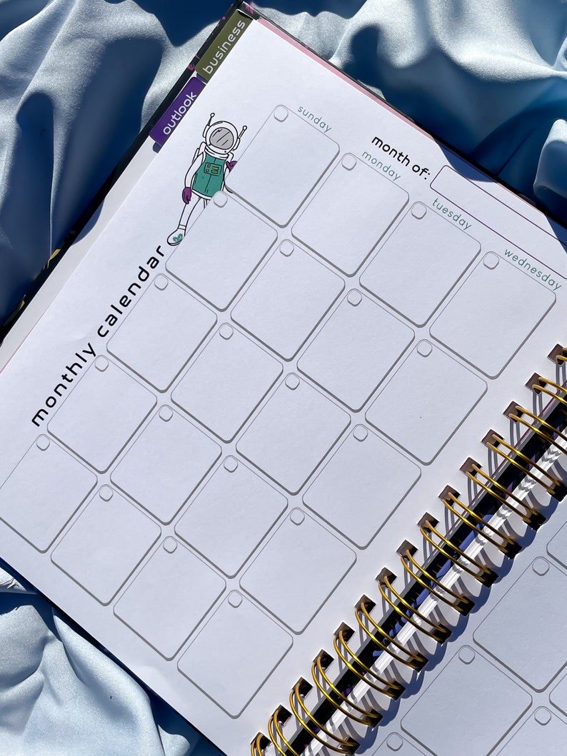 Pode incluir: Um calend&aacute;rio mensal com encaderna&ccedil;&atilde;o em espiral com interior branco e encaderna&ccedil;&atilde;o em metal dourado. O calend&aacute;rio apresenta um layout de grade com os dias da semana rotulados. Um desenho de astronauta est&aacute; no lado esquerdo. As palavras "calend&aacute;rio mensal" s&atilde;o impressas.