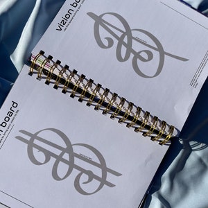 Pode incluir: Caderno espiral aberto com encaderna&ccedil;&atilde;o dourada. As p&aacute;ginas exibem um design de sinal de d&oacute;lar cinza e as palavras "vizion board" e "use this space to bring your vision to life". O caderno est&aacute; em tecido azul claro.